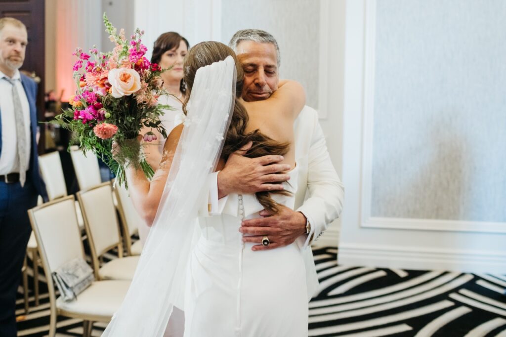 A bride in a white gown hugs her dad who is also in a white tux.