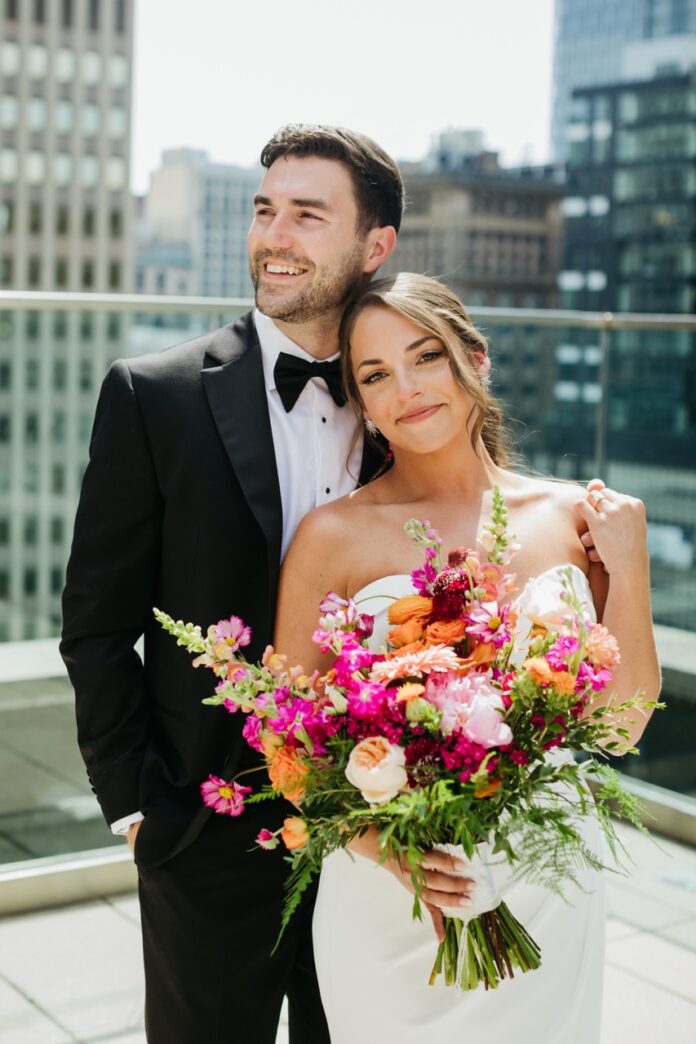 A bride in a white dress holds a colorful bouquet as she leans against her groom in a black tux.
