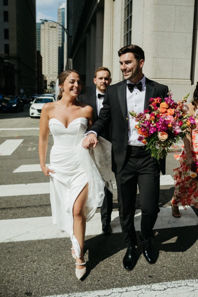 A bride in a white gown walks with a man in a black tux carrying a bouquet of flowers.