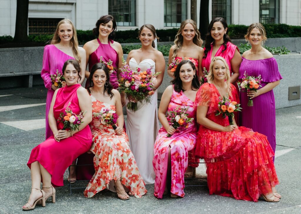 A group of bridesmaids in colorful dresses sit and stand around a bride in a white gown.