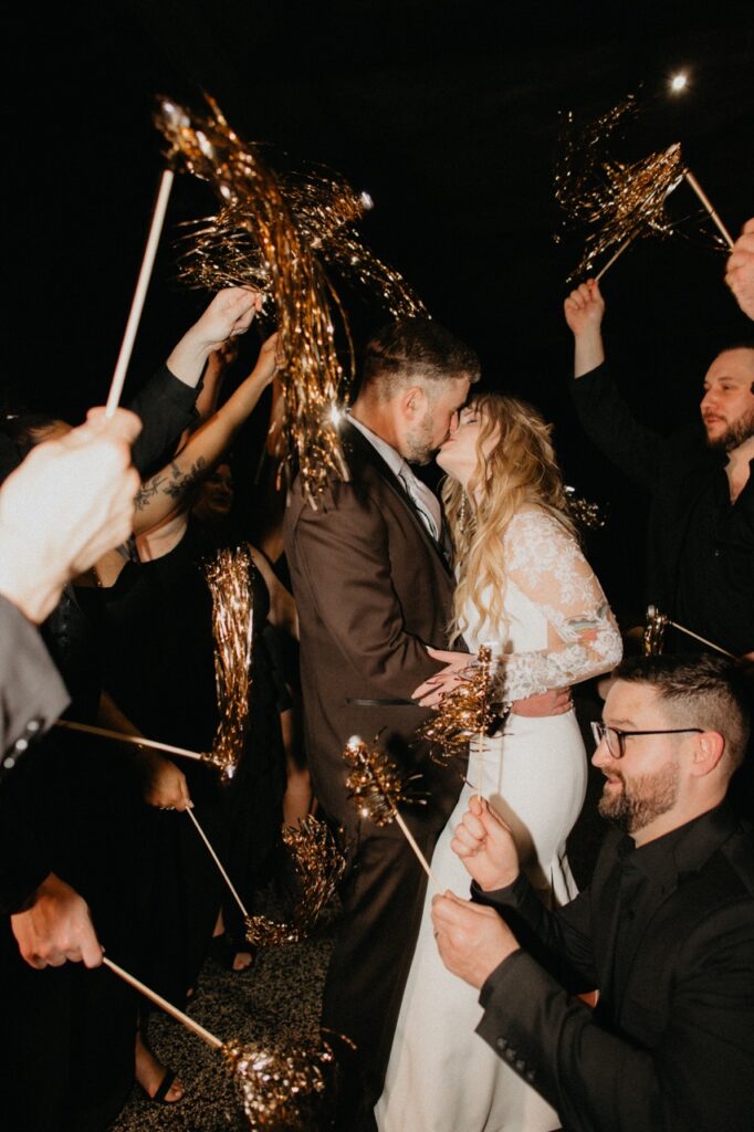 A bride in white and groom in black kiss behind gold streamers held by wedding guests.