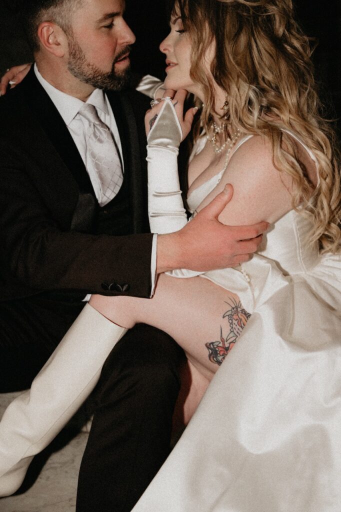 A bride in a white dress sits on a man in a black tux's lap.