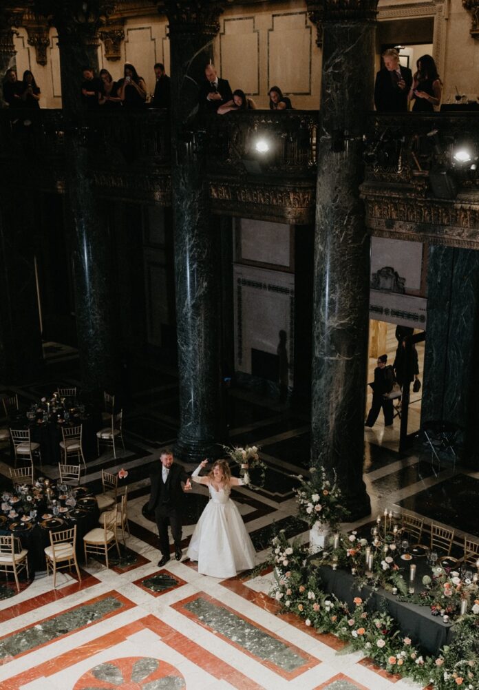 A bride in a white dress and groom in a black suit walk into a wedding reception.