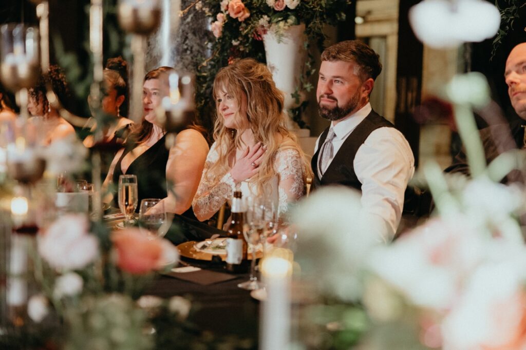 A woman in a white dress touches a hand to her heart as a man in dress clothes wraps an arm around her.