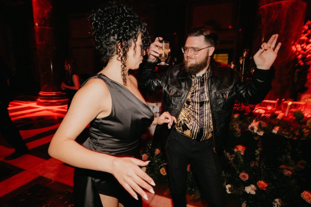 A woman in a black dress dances with a man in a black outfit with a drink in his hand.