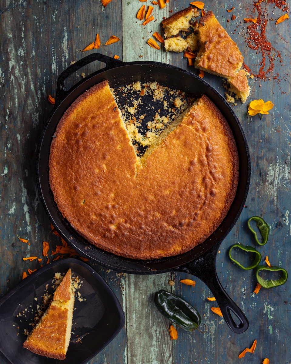 A cast iron pan of cornbread sits on a wooden table witha. slice cut out of it, sitting on a black plate nearby.