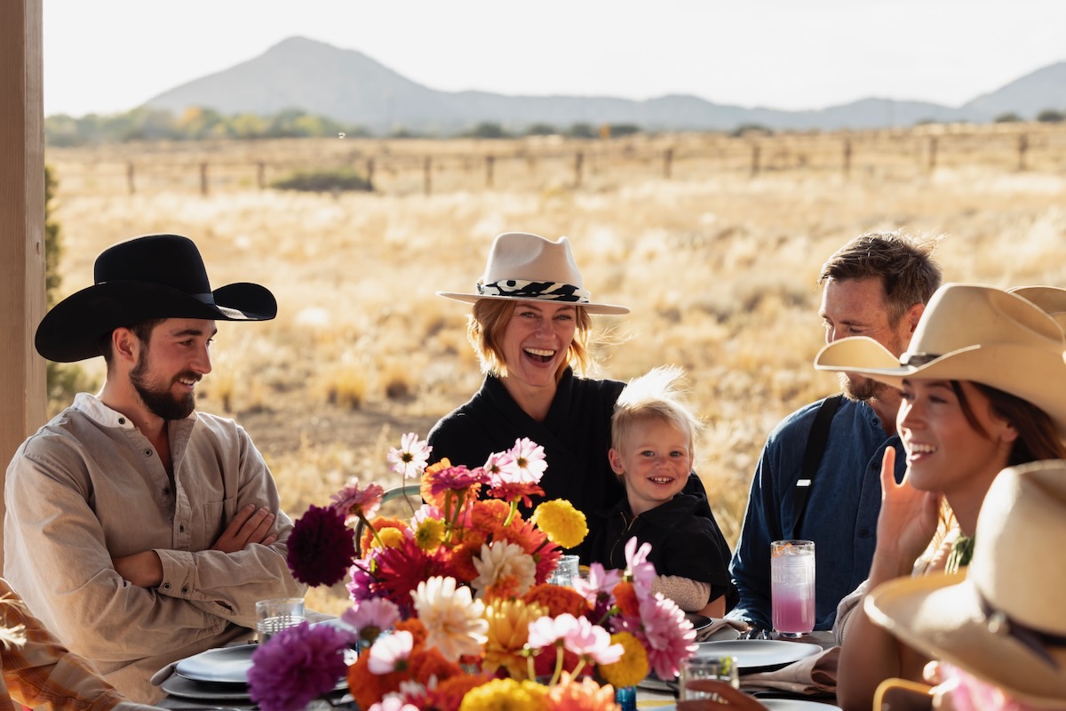 A woman in a cowboy hat and her child smile at the camera as two other men at the Double DD Ranch dinner party look at the table.