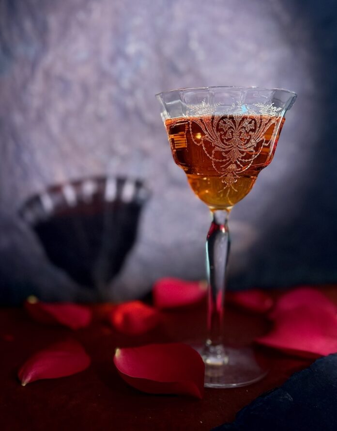 The Madama Butterfly Cocktail A Madama Butterfly inspired cocktail sits in a Nick and Norah glass against a purple background and rose petals scattered around the glass.