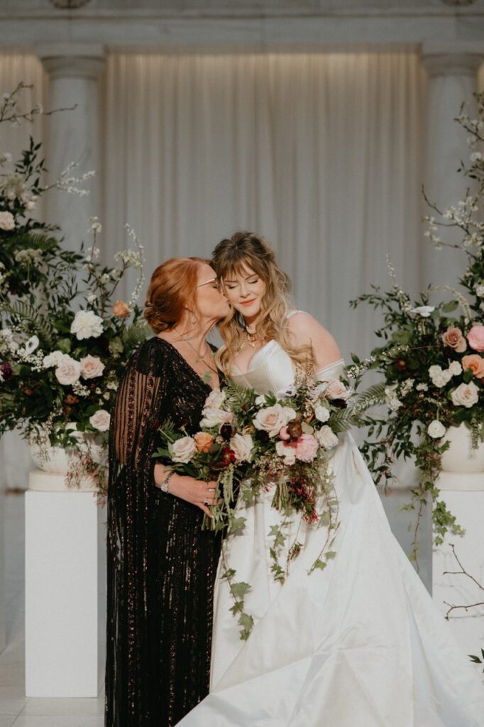 A bride in a white wedding gown, holding a huge bundle of flowers receives a kiss on the cheek from a woman in a black dress.