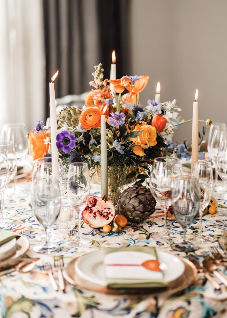 A table sits covered in white candles, orange and purple flowers, clear glasses, and white empty plates.