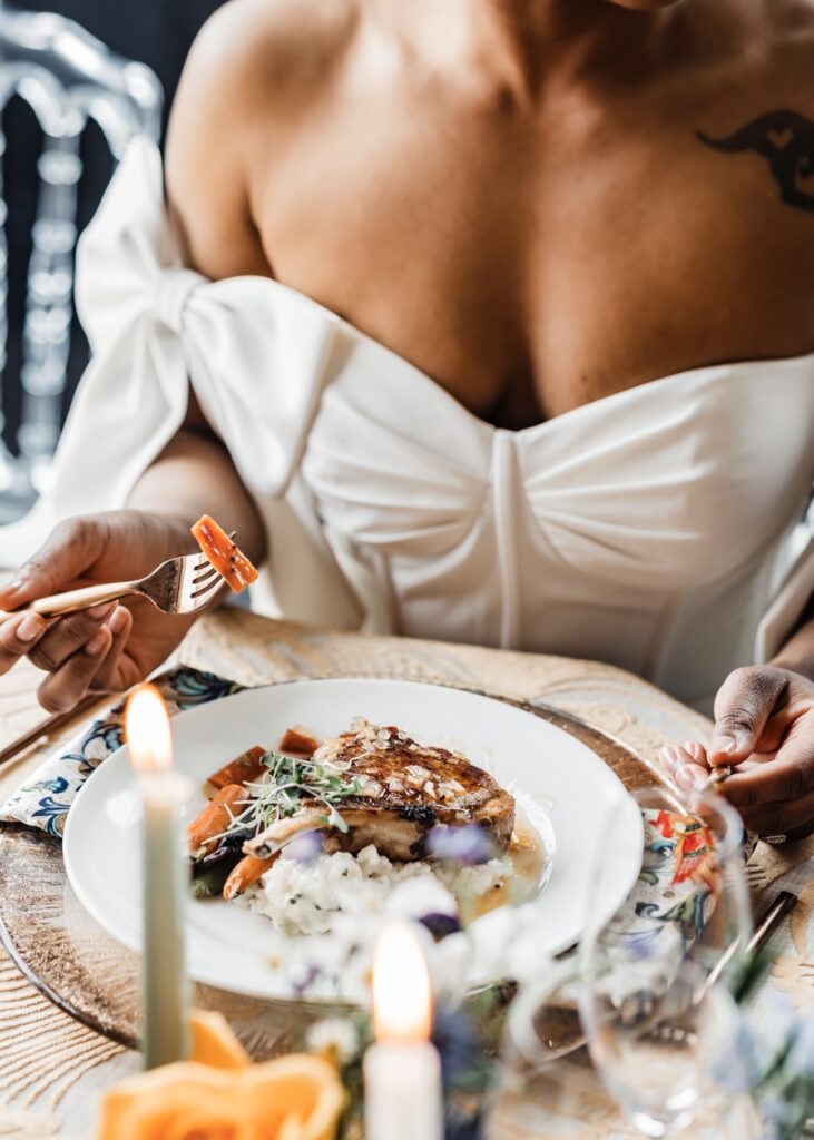 A bride in a white dress picks up a forkful of oranges off a white plate at her wedding.