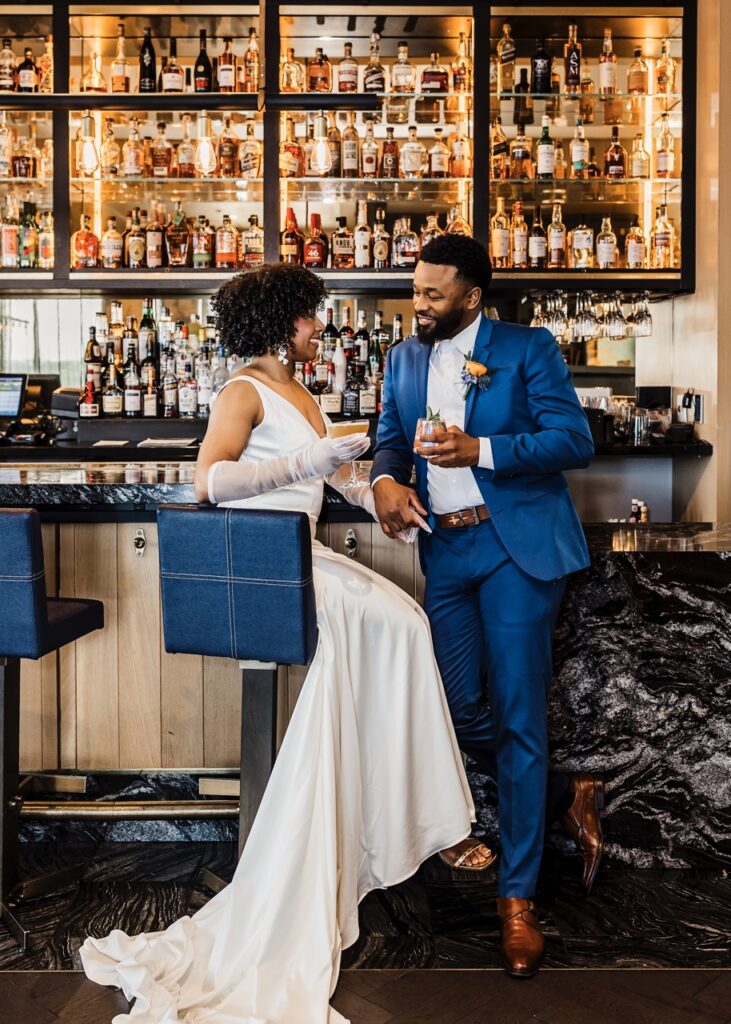 A bride in a white wedding gown sits in a blue chair, facing her groom who has a blue suit on.