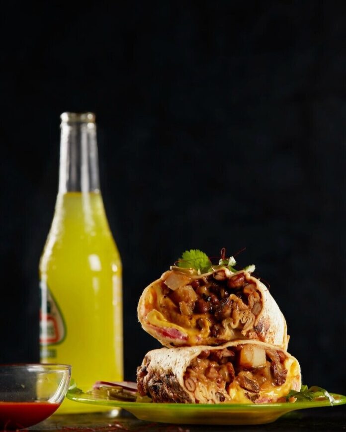 A breakfast burrito on a black background with a bottle of yellow juice.