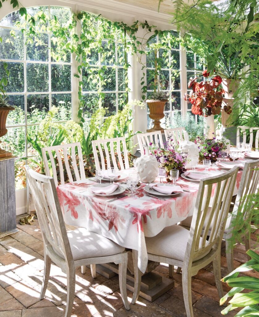 A dining room design by Bunny Williams using plenty of greenery, white chairs, and a pink and white tablecloth.