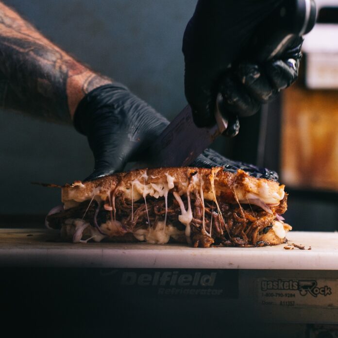 Cold Friends Kitchen is the Pittsburgh Food Truck You Need to Try A person wearing black gloves at Cold Friends Kitchen cuts a sandwich in half with a large knife.