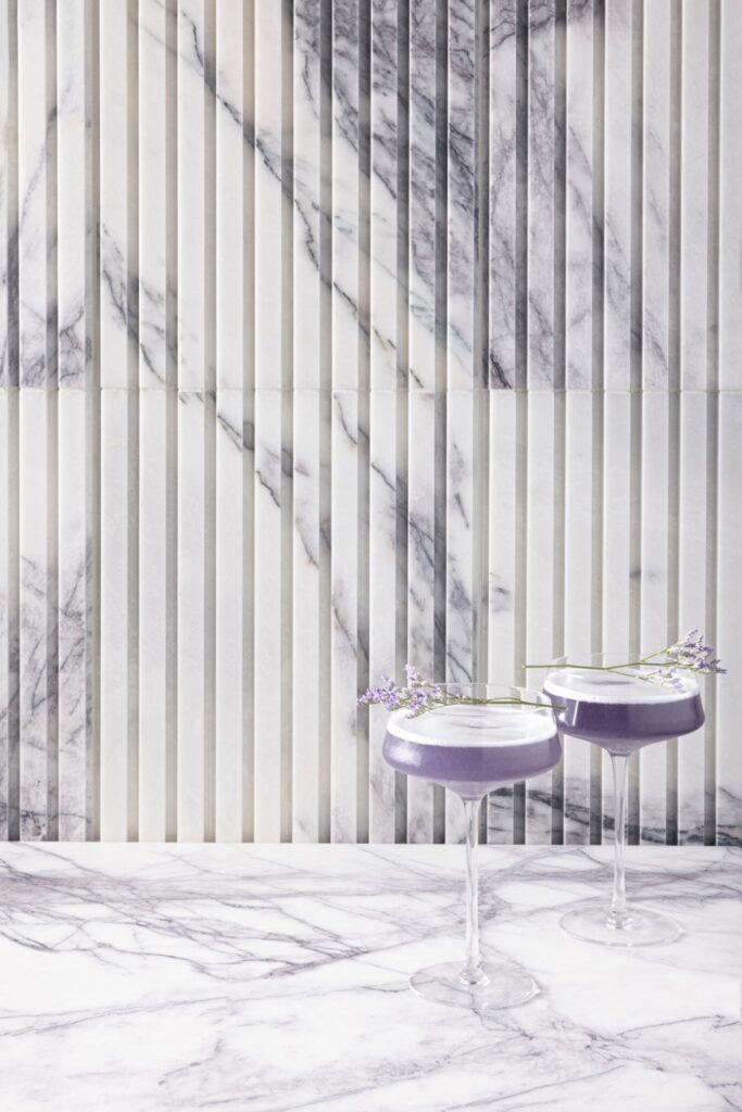 Two tall glasses hold surrealism purple cocktails against a lavender and white wallpaper.