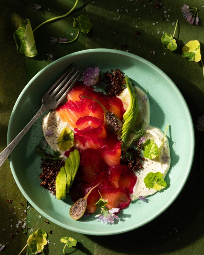 Thinly sliced salmon and beets to made a Beet Gravlax in a teal bowl surrounded by greens.