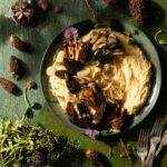 A green bowl on a green table holds cheesy grits inside mixed with small blossoms and springtime mushrooms with uncooked mushrooms all around the bowl.