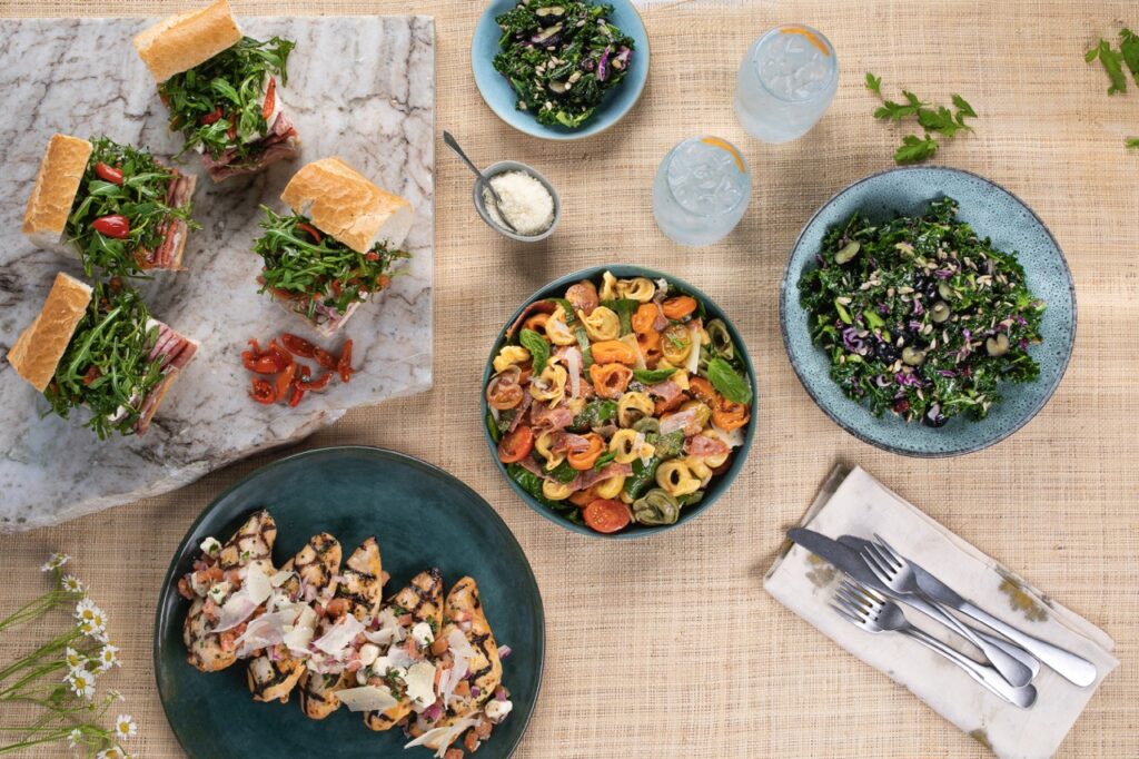 Various catering dishes in bowls and plates sit on a brown table with little sandwiches to the left and salads to the right.