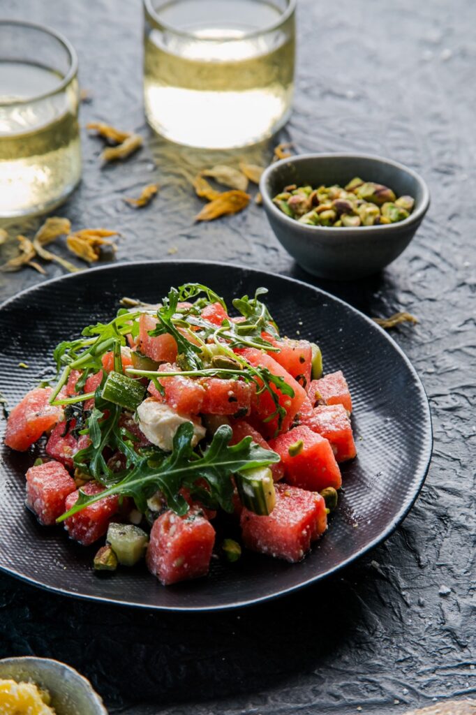 A watermelon salad from Common Plea Catering sits on a dark blue plate with a bowl of seeds nearby.