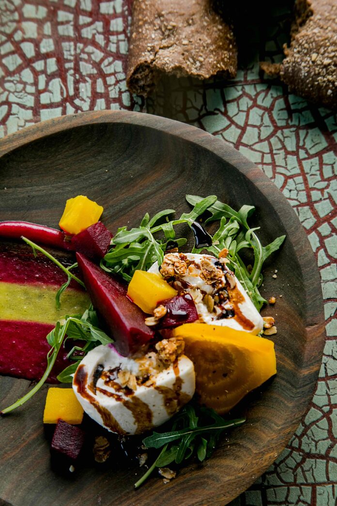 A wooden dish sitting on a crackle-finished surface with yellow and red beets, burrata and arugula, drizzled with balsamic vinegar made by Common Plea Catering.