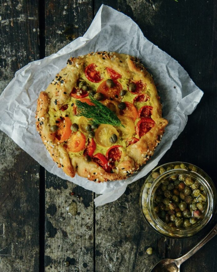 An Everything Bagel Tomato Tartelette sits on a piece of parchment paper with a jar of capers sitting nearby.