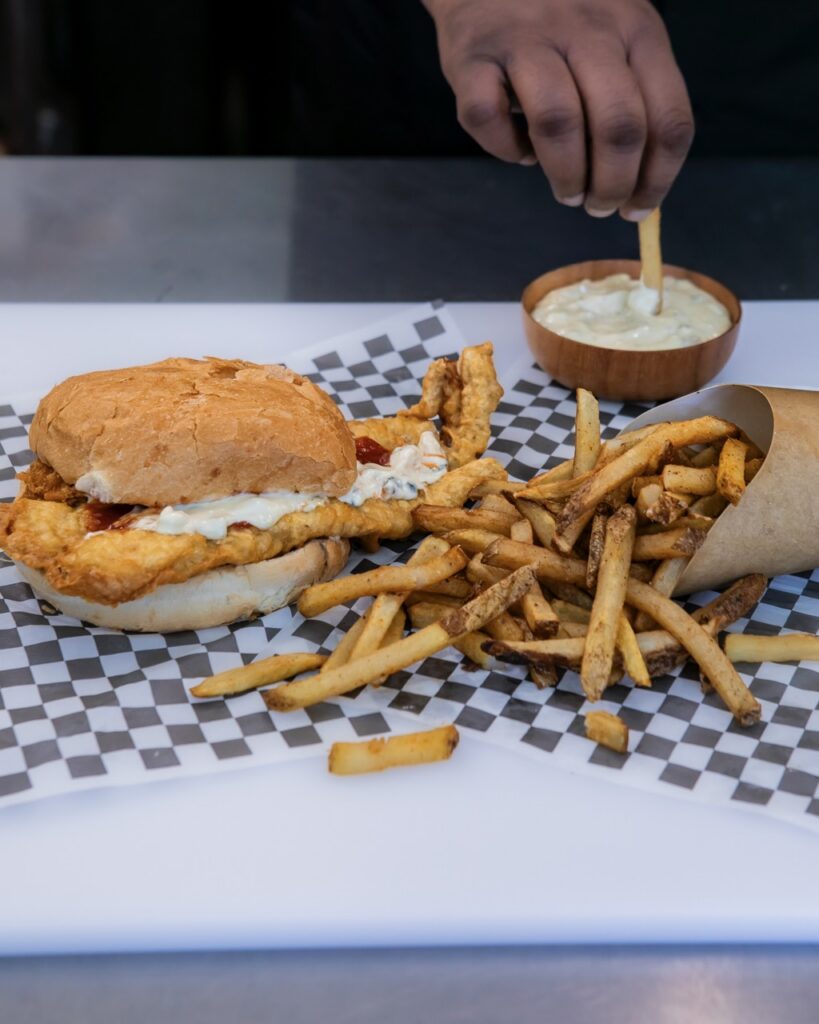 A huge dish sandwich beside fries that a man dips in tartar sauce.