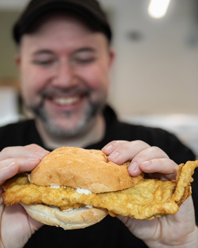 A man holds a fried fish sandwich with white sauce.