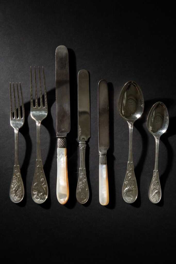 Two forks, three knives, and two spoons from the Frick's gilded tableware collection sit in a row on a black tabletop.