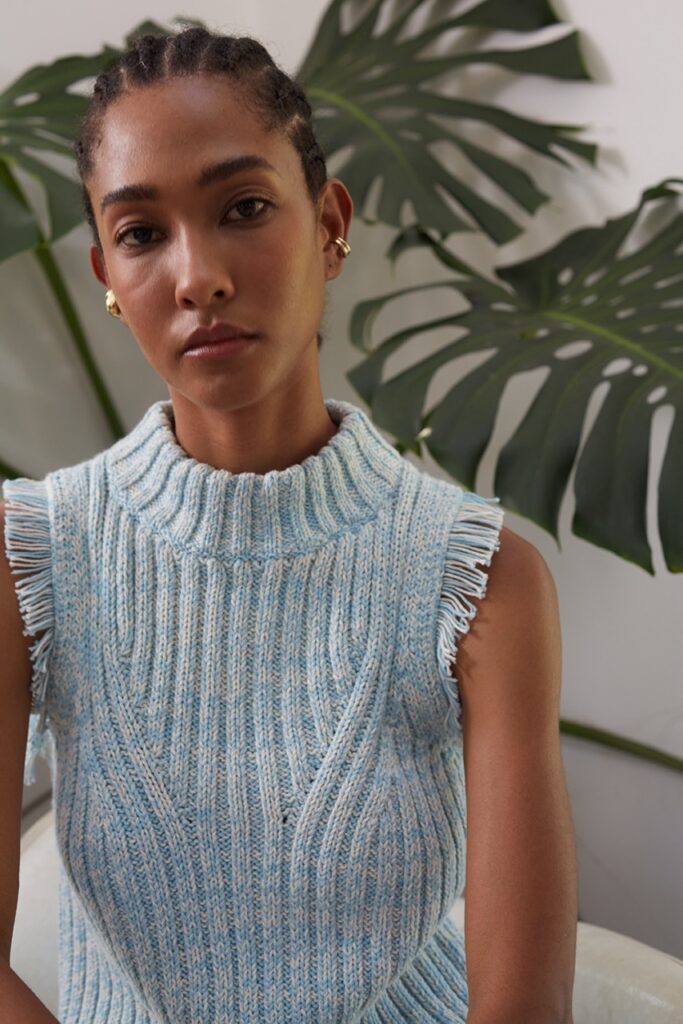 A woman stands in front of a houseplant in a blue sweater vest.