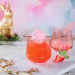 A short wine glass with a Here Comes Peter Cottontail inside as a ball of cotton candy sits over the top of the glass. A golden bunny sits in the background by a pastel backdrop.