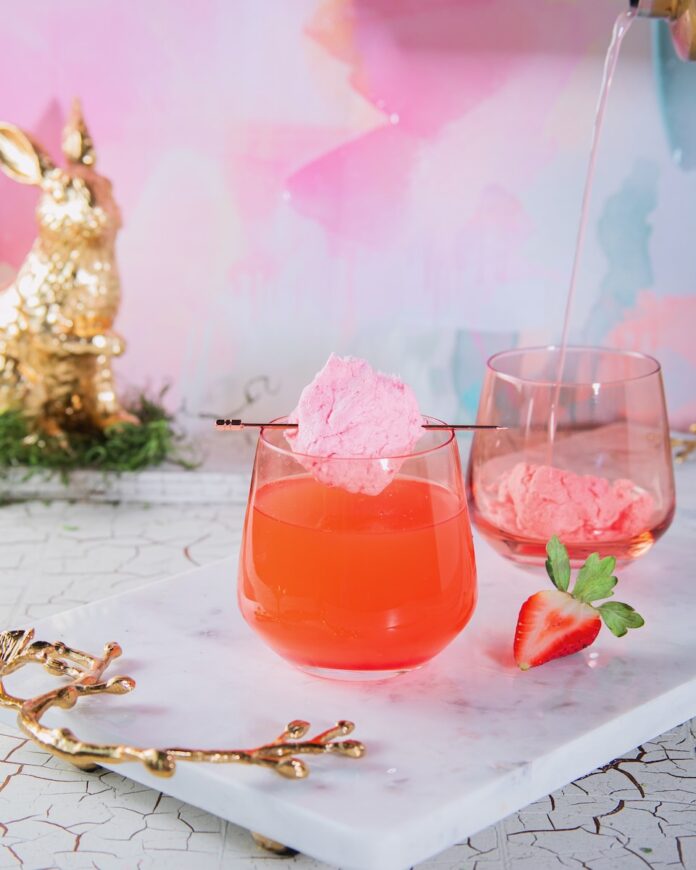 Here Comes Peter Cottontail Cocktail A short wine glass with a Here Comes Peter Cottontail inside as a ball of cotton candy sits over the top of the glass. A golden bunny sits in the background by a pastel backdrop.