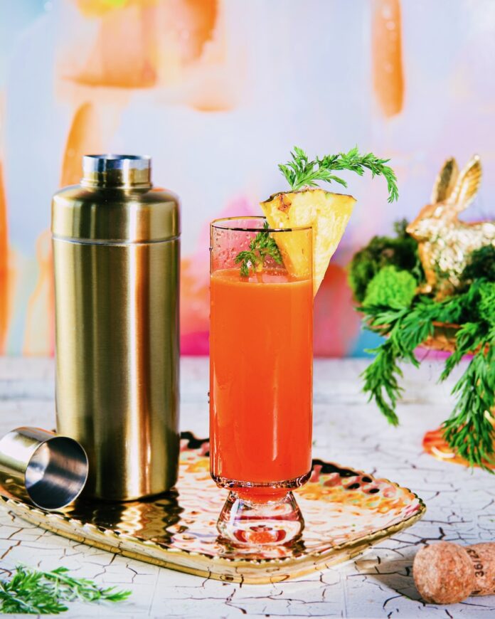 Hoppin’ Down the Bunny Trail with a Buzzed Bunny Mimosa A tall glass holds a Hoppin’ Down the Bunny Trail with a Buzzed Bunny Mimosa in an orange color with a pineapple slice garnish as the whole glass sits beside a gold cocktail shaker.