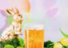 Peter Cottontail’s Easter Cocktails and Mocktails A Hoppity IPA Cocktail in a tall beer glass sits on a white table surrounded by Easter grass, eggs, and a gold bunny by the pastel backdrop.