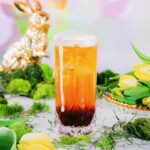 A Hoppity IPA Cocktail in a tall beer glass sits on a white table surrounded by Easter grass, eggs, and a gold bunny by the pastel backdrop.