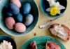 Italian Easter Bread On a green plate beside a bowl of brightly colored easter eggs sits a round braided Italian Easter Bread with a slice taken out of the top which sits on the table buttered.