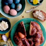 On a green plate beside a bowl of brightly colored easter eggs sits a round braided Italian Easter Bread with a slice taken out of the top which sits on the table buttered.