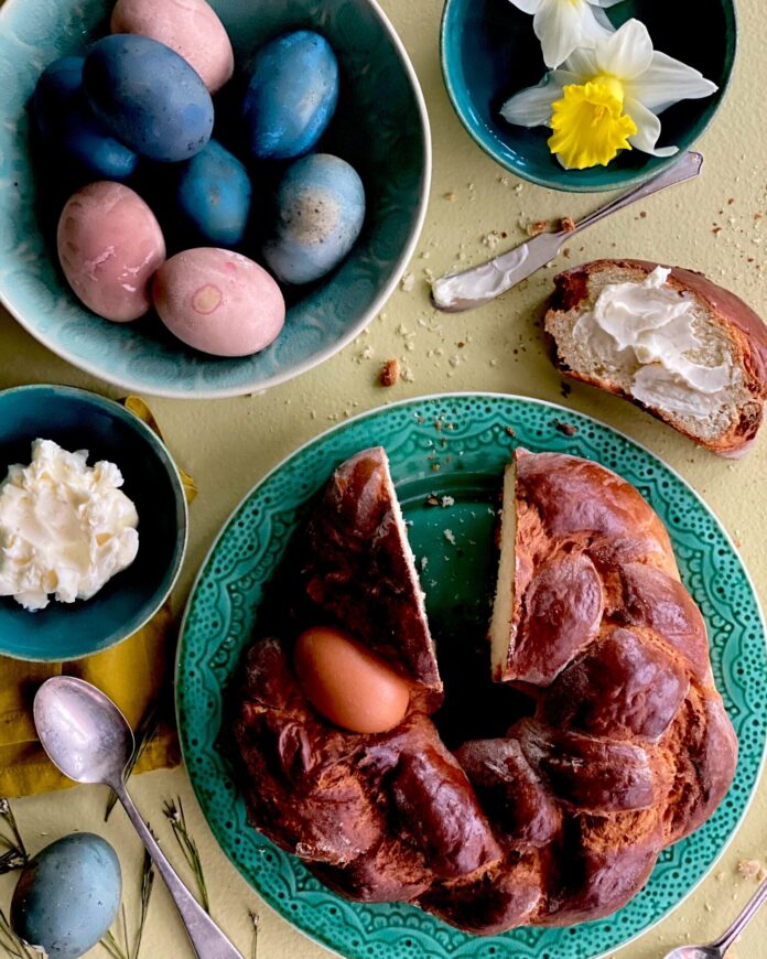 On a green plate beside a bowl of brightly colored easter eggs sits a round braided Italian Easter Bread with a slice taken out of the top which sits on the table buttered.