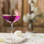 A coupe glass with a purple Mother's Day Gin and Tonic mocktail with crushed freeze-dried blueberries on the side and an edible pink butterfly on the rim. White flowers are scattered nearby.