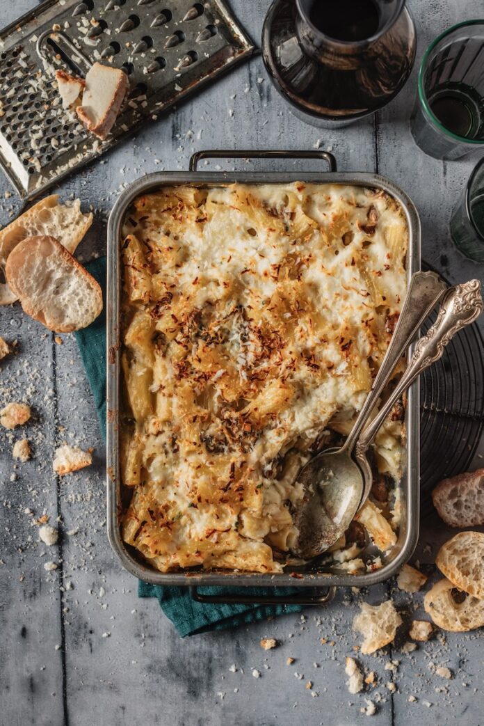 Pasta au Gratin A baking dish with crusty bread sitting around it on a grey table holds a Pasta au Gratin with a spoon scooping out the bottom right corner.