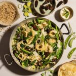In a large steel pot sits a Pea Pasta Primavera with peas and other ingredients surrounding the pot on a white table.