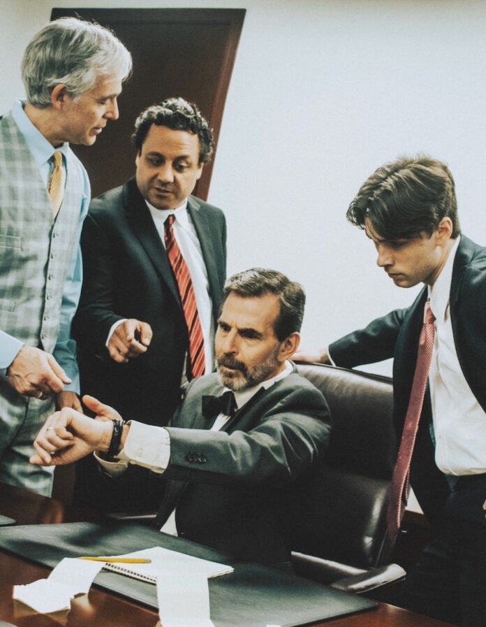 Pittsburgh Events: Your Guide to April 28-May 4, 2025 Three men in a Pittsburgh play surround a man seated at a desk as they all look at his watch.