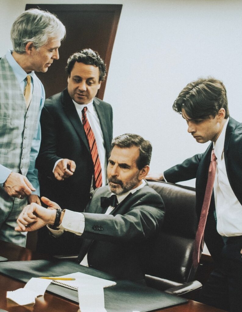 Three men in a Pittsburgh play surround a man seated at a desk as they all look at his watch.