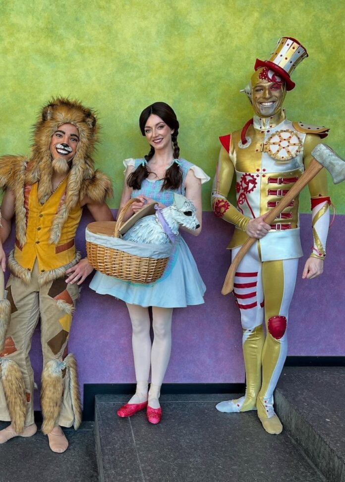Pittsburgh Events Wizard of Oz Ballet TABLE Magazine Dorthy, the lion, and the tin man stand in Pittsburgh ballet outfits against a green and purple background.