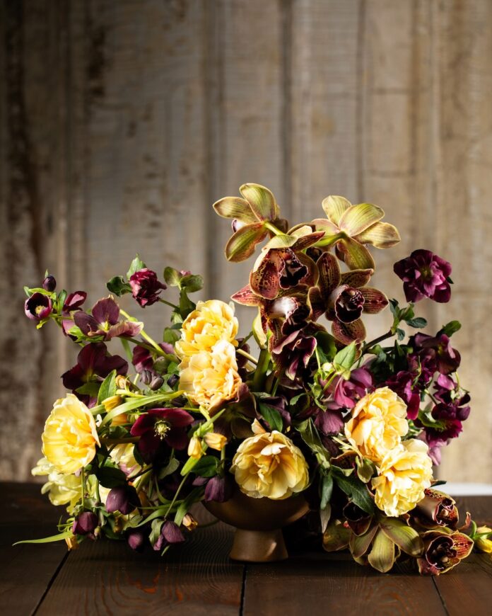 A floral arrangement by a Pittsburgh florist sits on a table in yellow and burgundy blooms with a wood panel wall behind it.