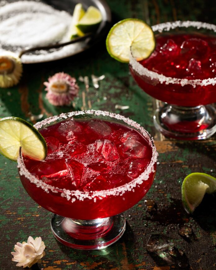 Pomegranate Margarita Two margarita glasses with salt rims are filled with a red pomegranate margarita with a lime wheel garnishing each glass.