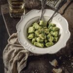 A white bowl holds Potato and Pea Gnocchi in a green color with two forks sitting beside the bowl and bread sitting above it.