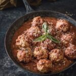 A bunch of ricotta vegetarian meatballs sit in a cast iron pan, soaked in re sauce with basil garnish on top as the pan is surrounded by pasta, a bowl of the meatballs, and bread.
