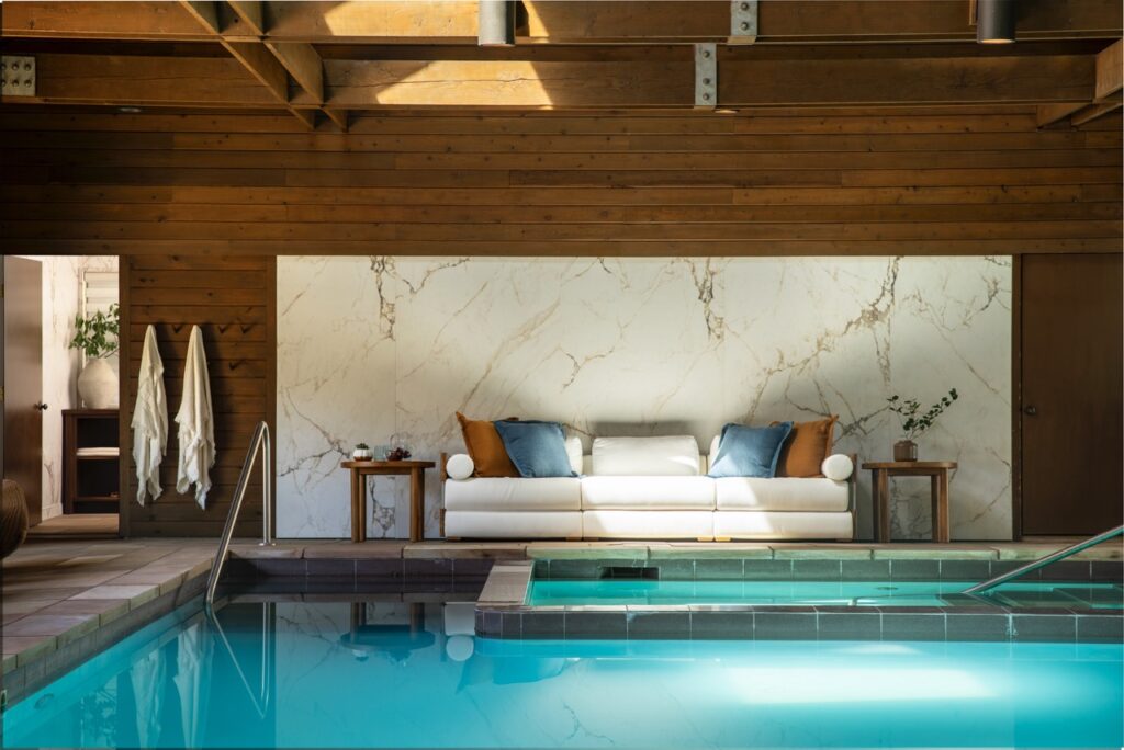 An indoor pool with blue water sits below a small white couch against a marble wall.