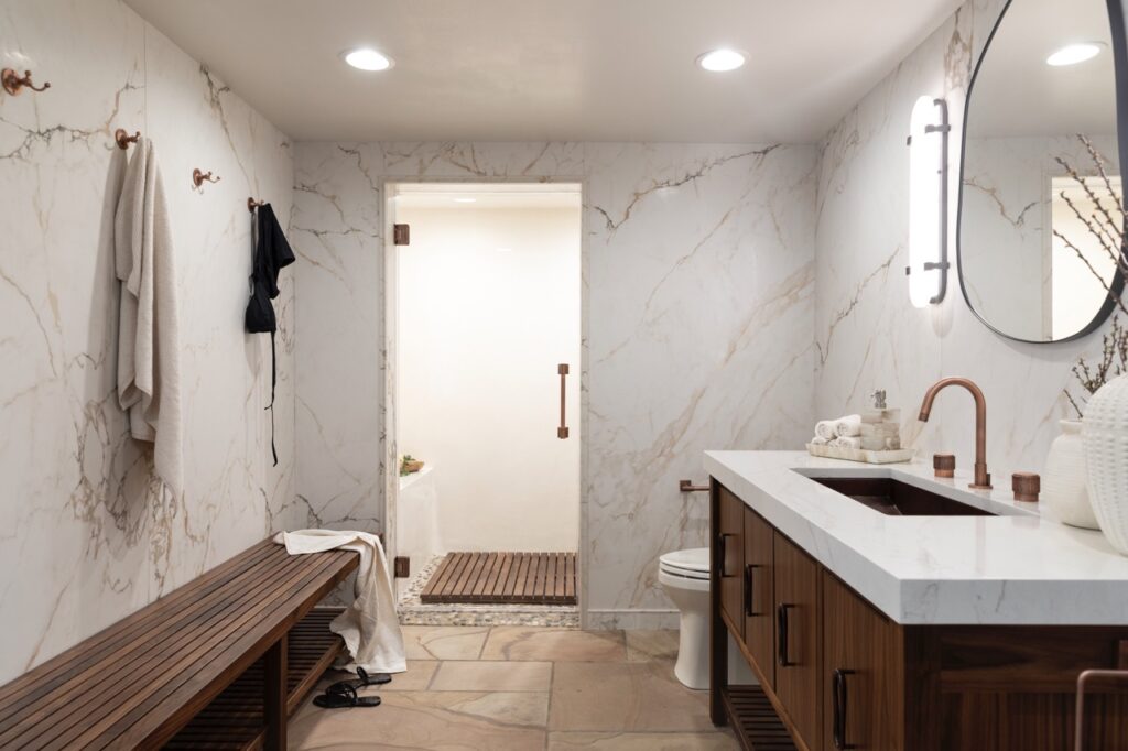 A bathroom designed by Seashal Belldina with white marble walls, dark wood finishes, a sink, a mirror, and a doorway.